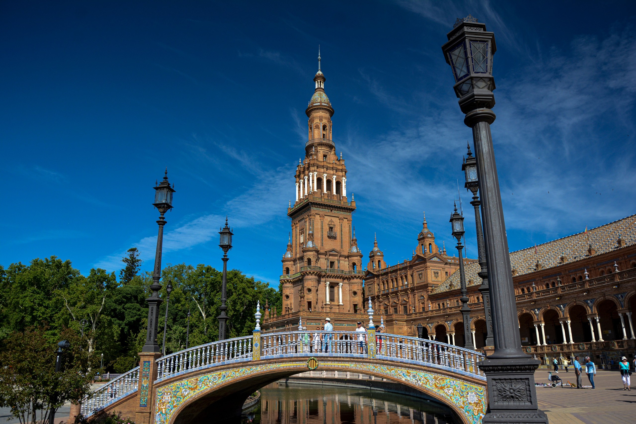 People walking on tiled bridge near tall brick tower with ornate design under blue sky.