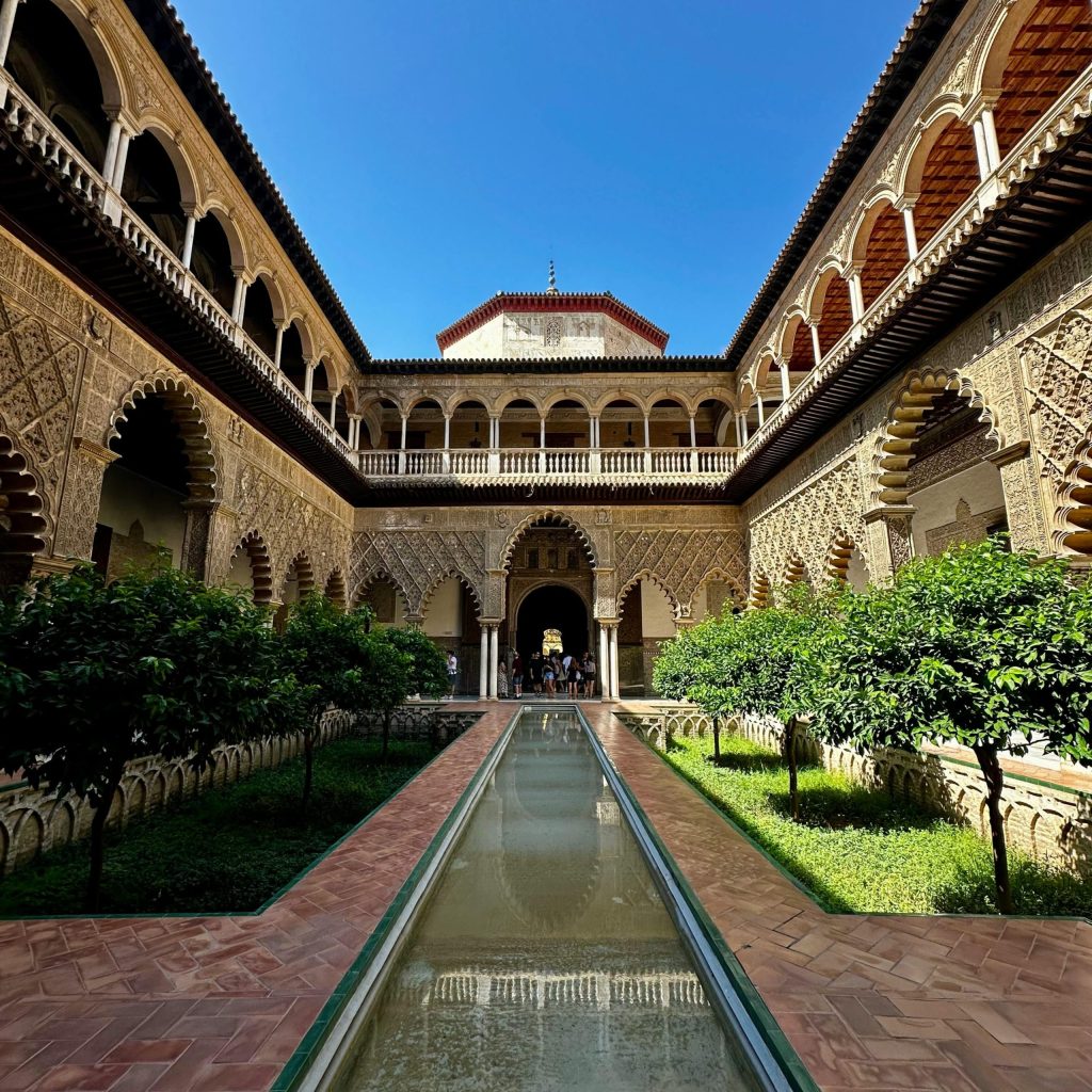 Courtyard with ornate arches, central pool, and green trees under a clear blue sky.