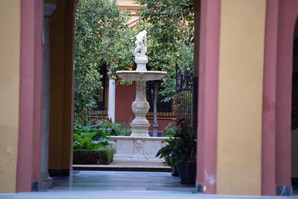 View of a courtyard with a fountain and orange trees through an archway.