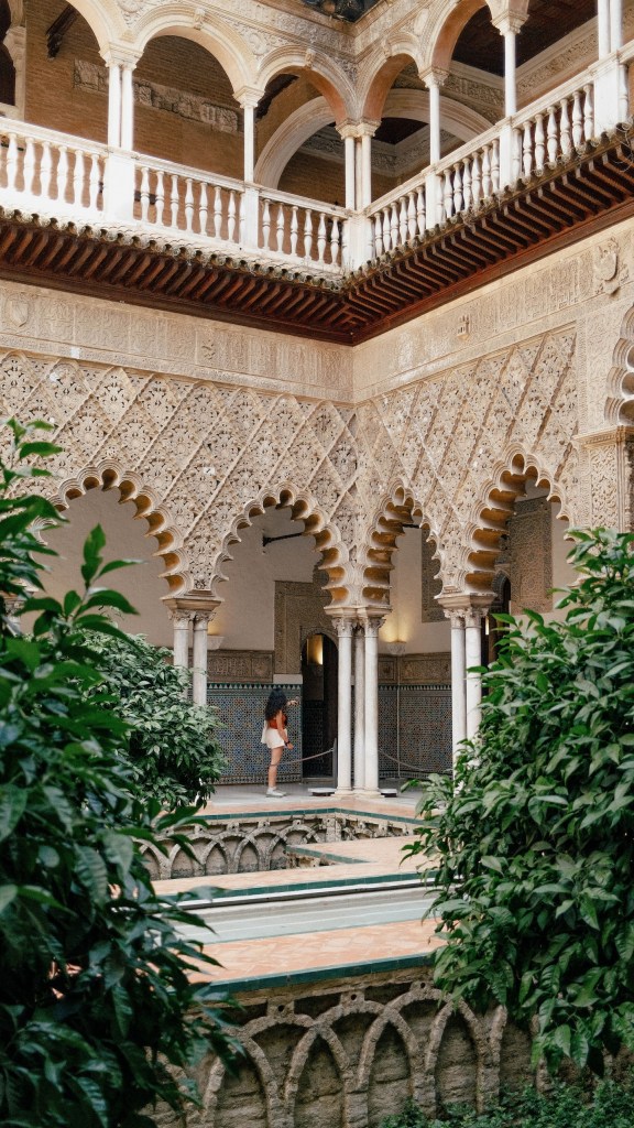 Person standing in ornate, arched courtyard with lush greenery.