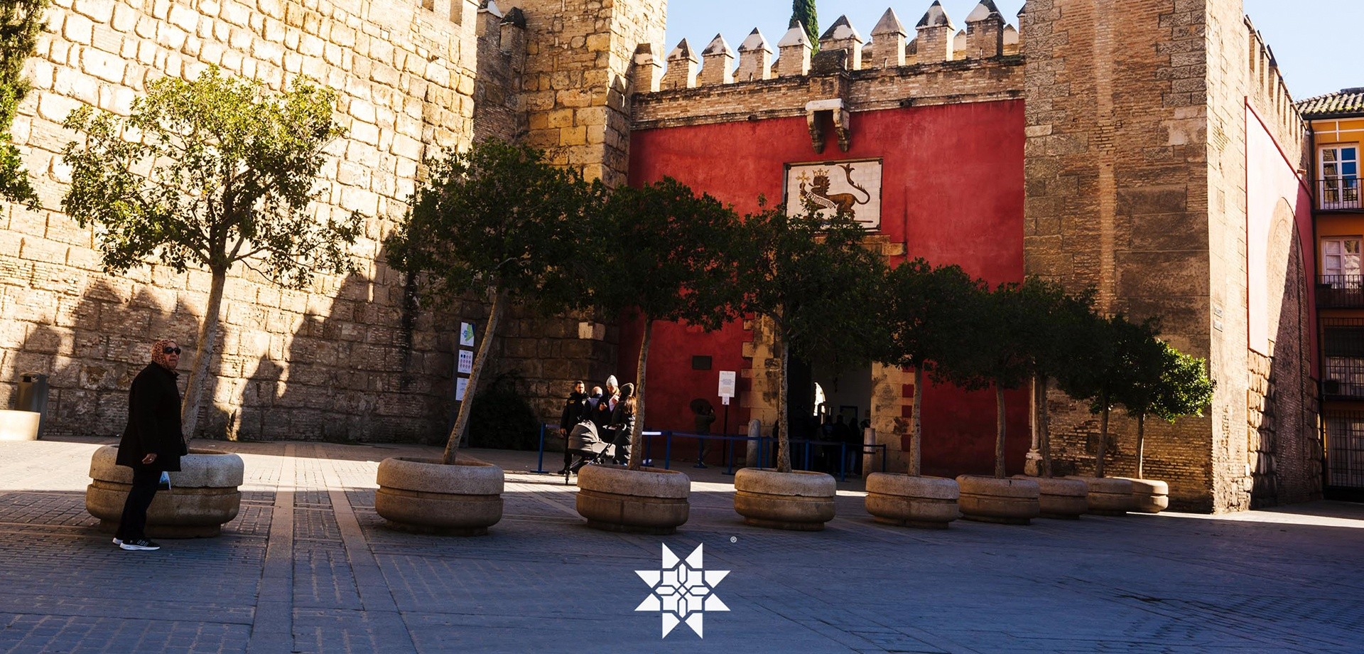 Historic building with red walls, trees, and people in a sunny plaza.