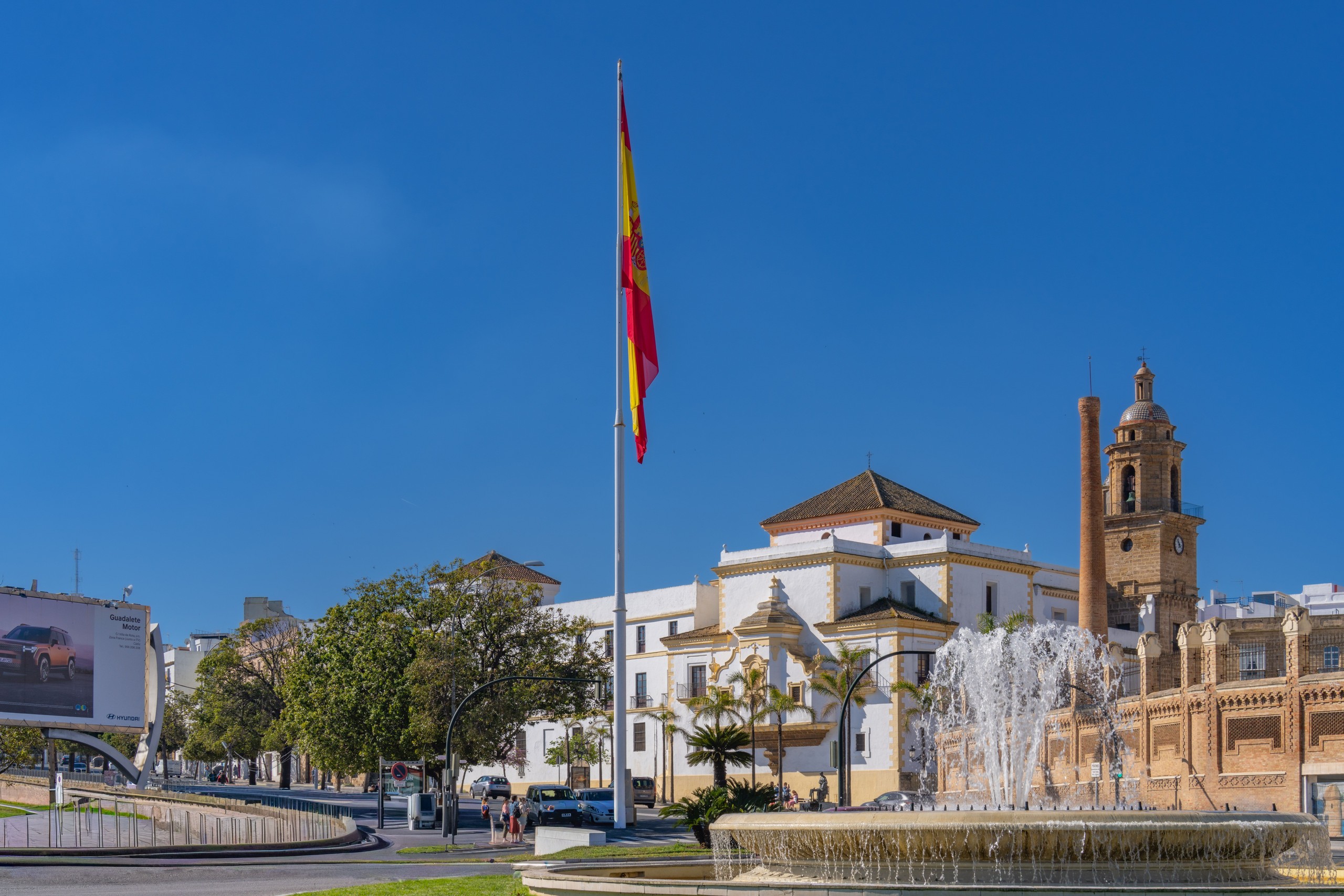 Spanish flag near a fountain and historic buildings under a clear blue sky.