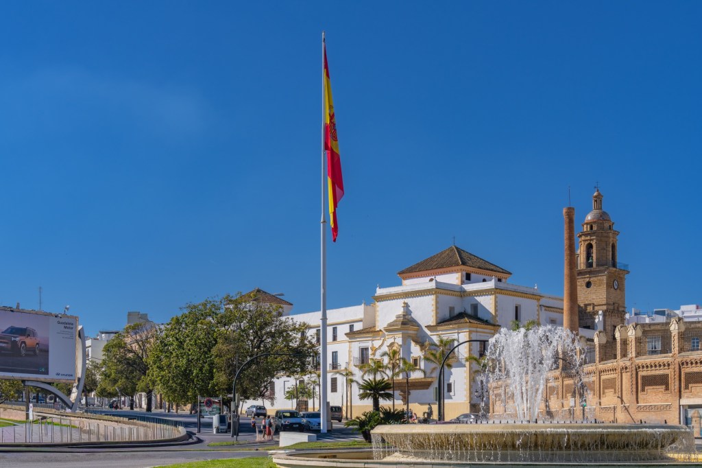 Spanish flag near a fountain and historic buildings under a clear blue sky.