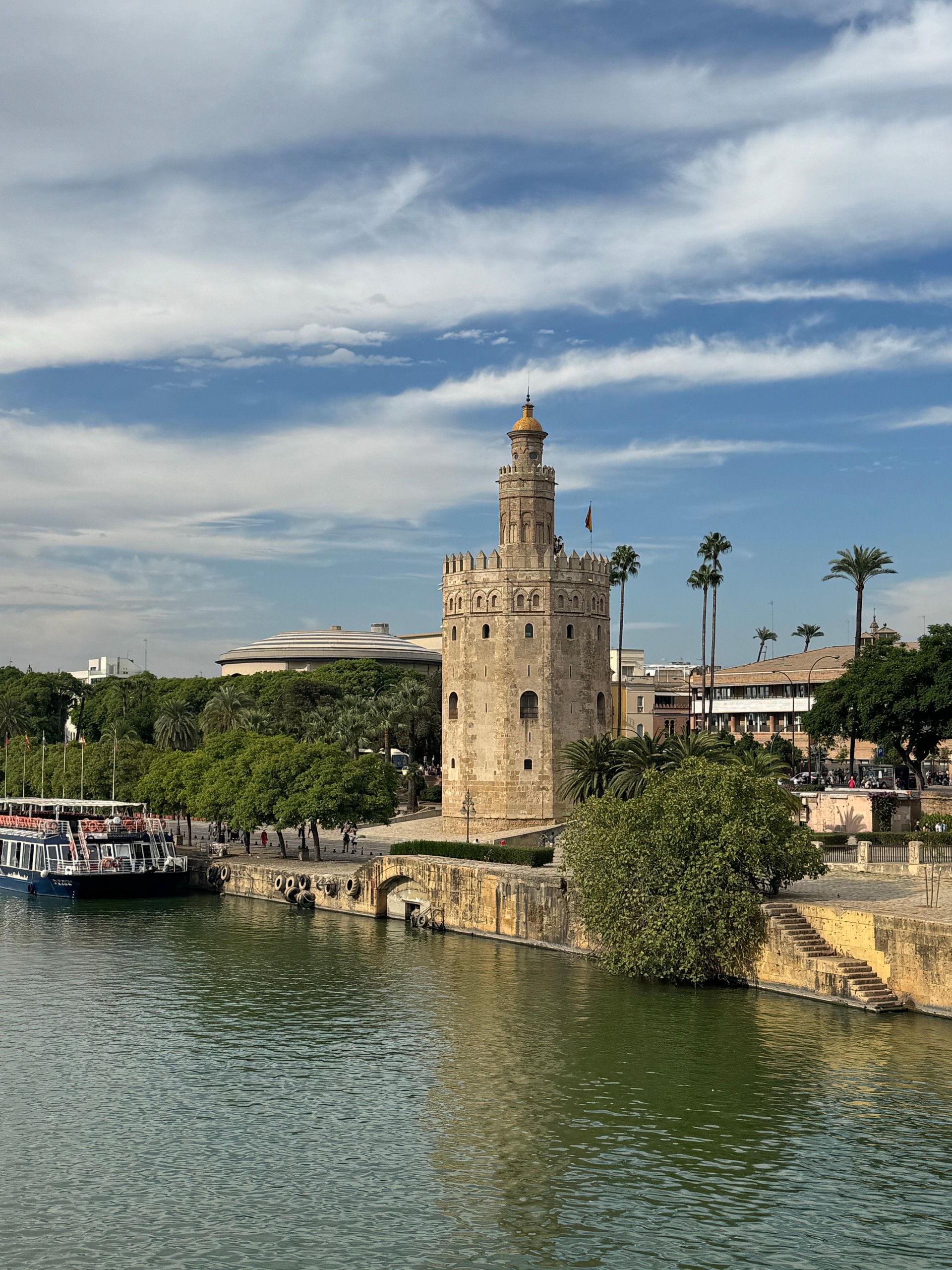 Historic stone tower by a river with boats and palm trees on a sunny day.