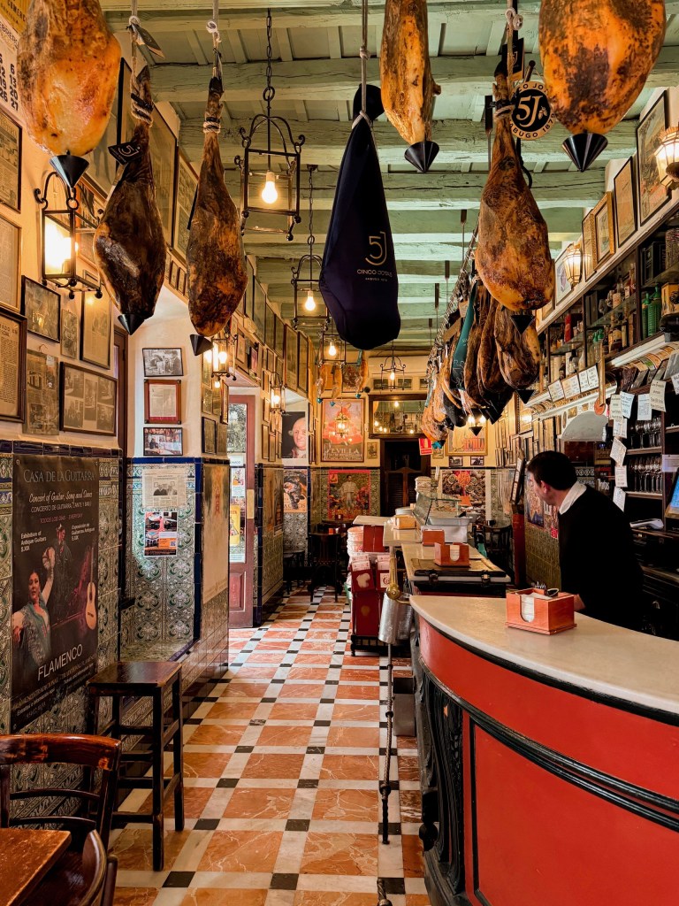 Cozy tapas bar with hanging hams, tiled floor, and a curved bar counter.