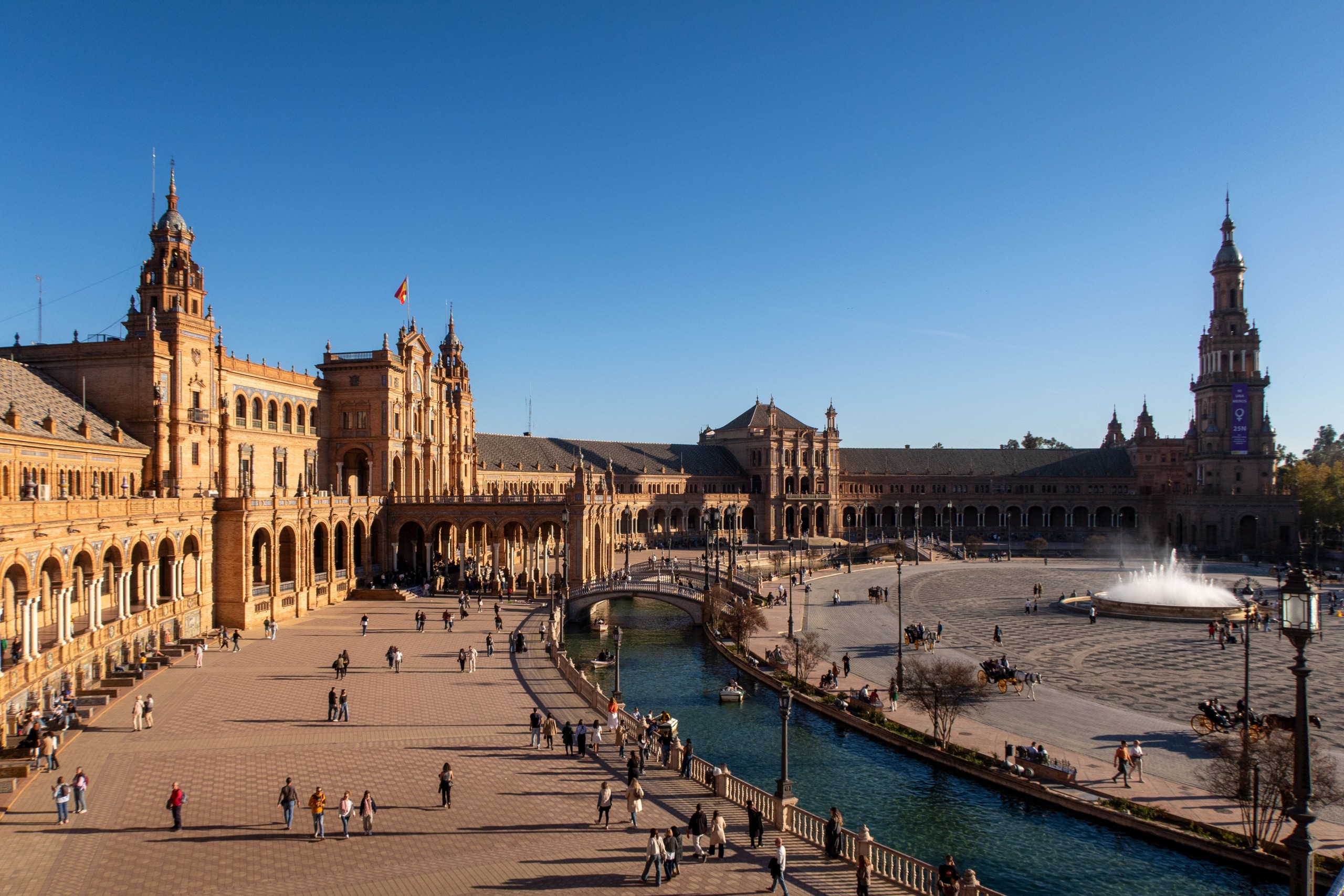 Crowded Plaza with historic buildings, tower, canal, and fountain on a sunny day.